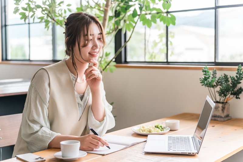 ノートPCの前でノートに書き込みながら作業する女性