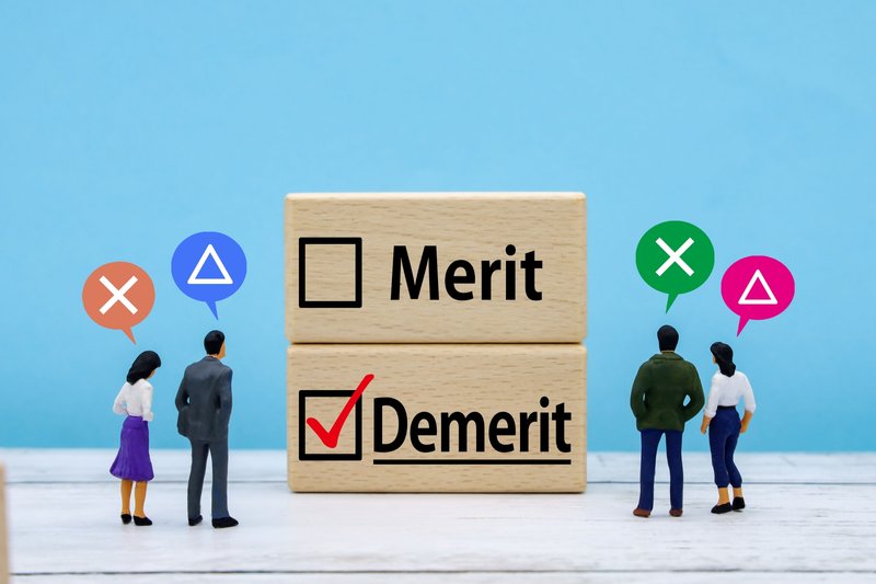 Merit・Demeritと書かれた積み木の前でミニチュア人形が議論しているコンセプト写真